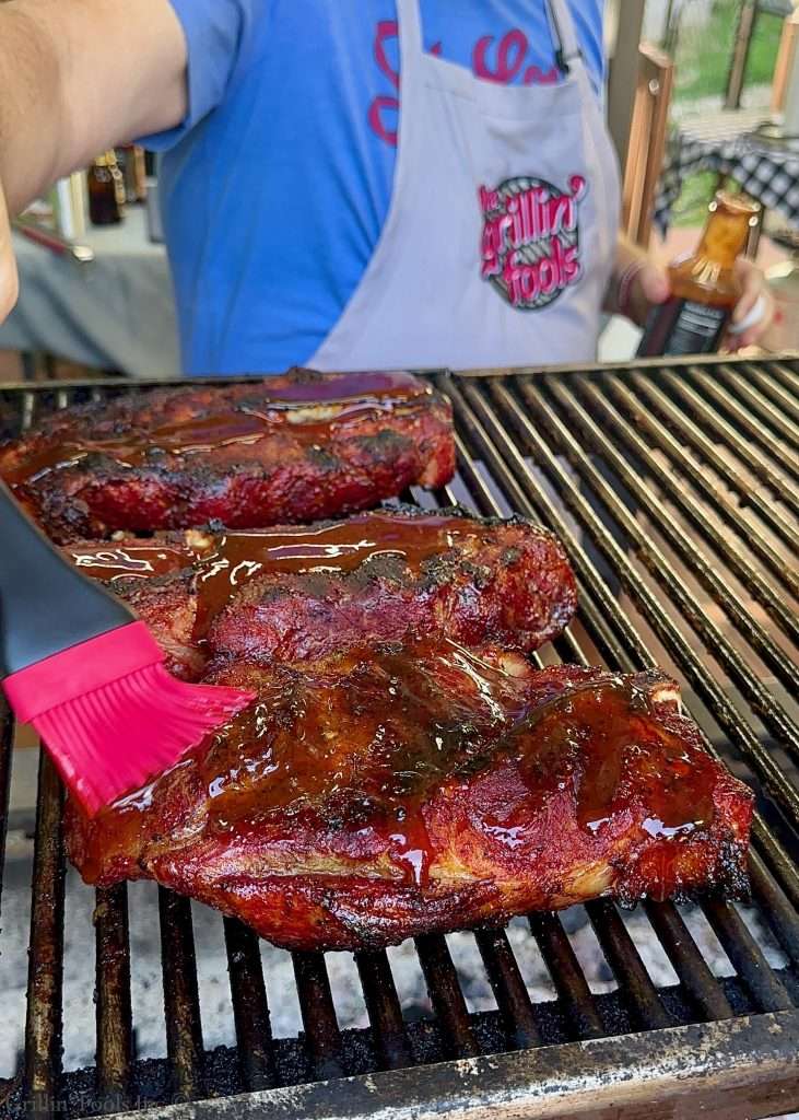 Barbecued pork steaks on a grill; a person in a blue shirt and grey 'GrillinFools.com' apron appears to apply sauce with a red basting brush.