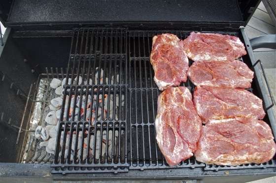 Raw pork steaks arranged on the right side of a grill with hot charcoal on the left for a two-zone grilling set up.