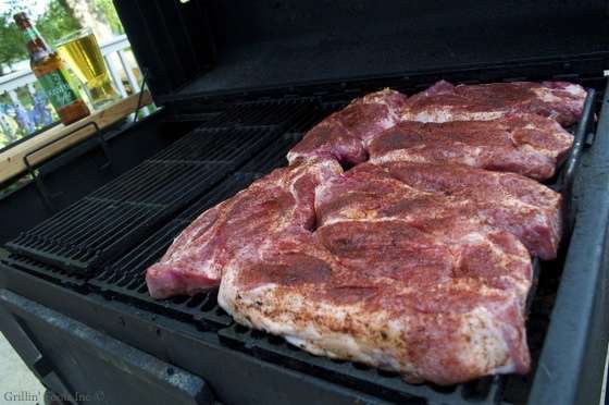 Large slabs of seasoned raw pork ribs laid on the near side of a barbecue grill outdoors, with bottle and glass of beer visible on a table in the background.