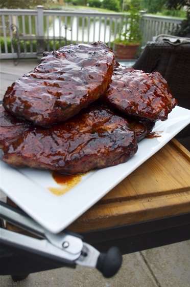 Stack of glossy barbecue pork steaks stacked on a white rectangular platter outdoors on a wooden table with a lake in the background