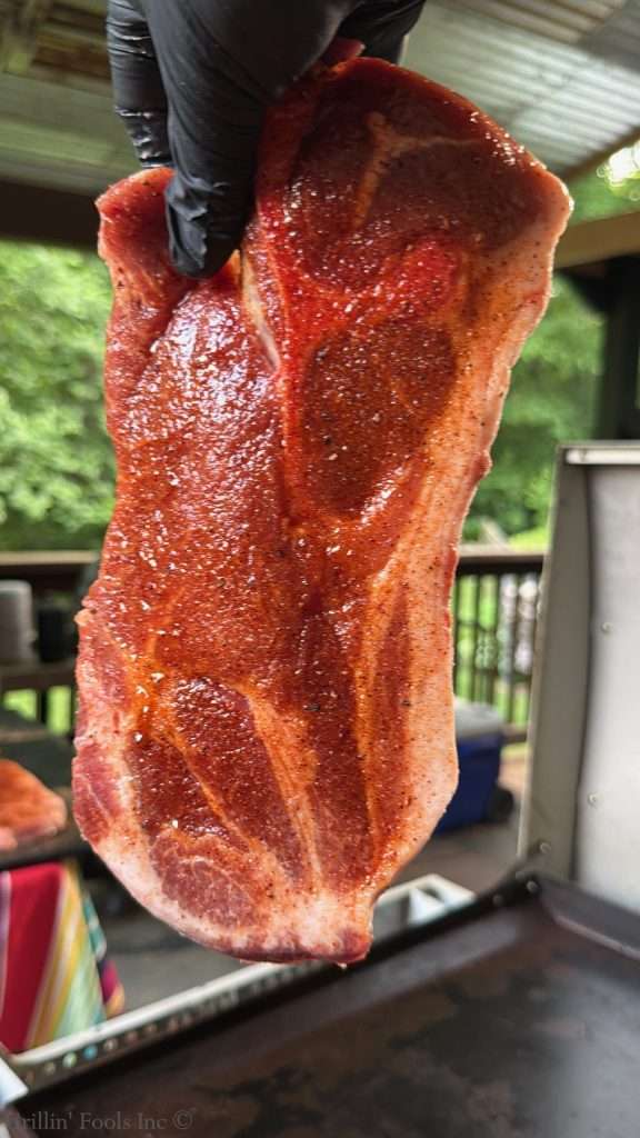 A raw, seasoned pork steak being held by a gloved hand outdoors over a flat top griddle, ready for grilling.