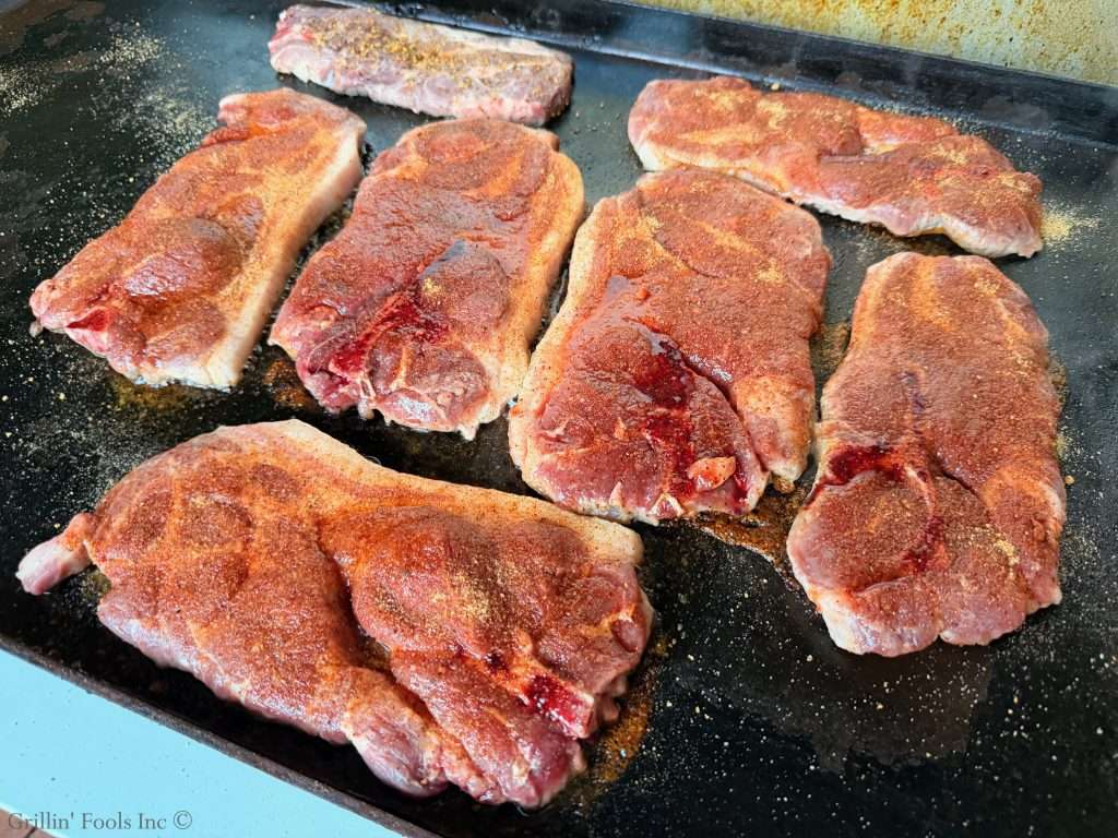 Several raw, bone-in pork steaks seasoned with a reddish spice blend on a hot griddle