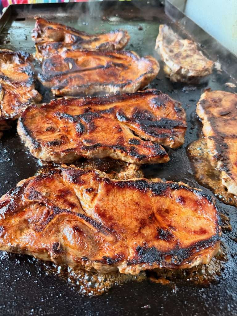 Several pork steaks cooking on a flat grill with dark, crispy crusts and sizzling juices nearby.