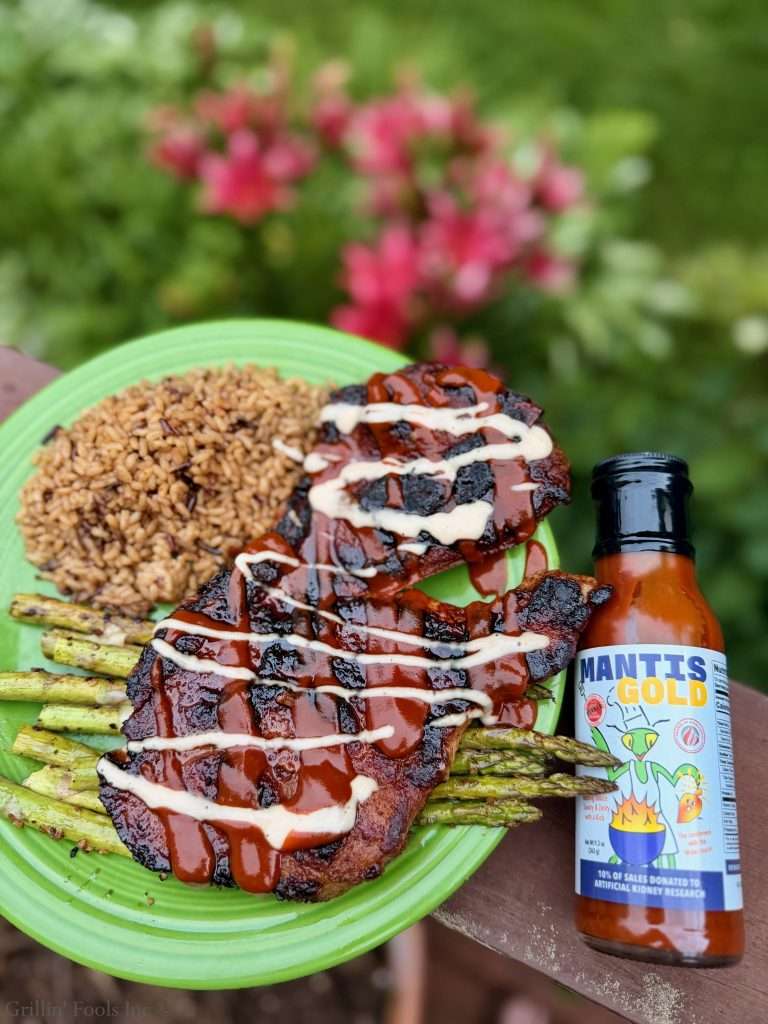 Barbecue pork steak with Mantis Gold BBQ sauce and a drizzle of Alabama white sauce on a green plate, with brown rice and grilled asparagus, beside a Mantis Gold sauce bottle.
