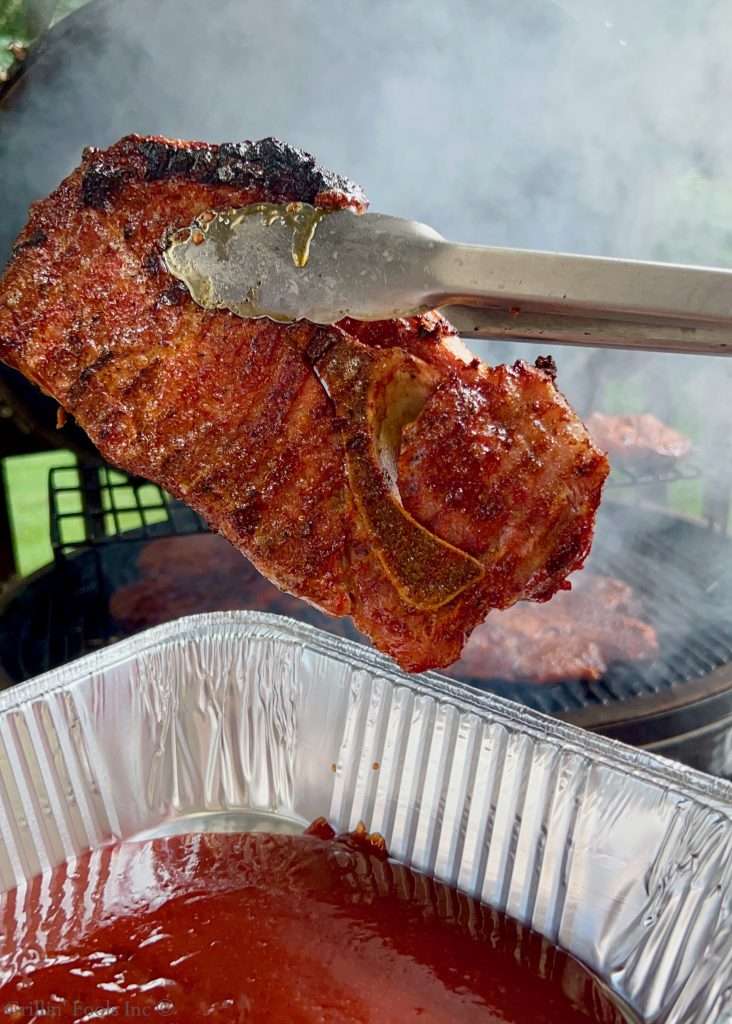 Close-up of a smoked pork steak being sauced with tongs over a smoky grill.