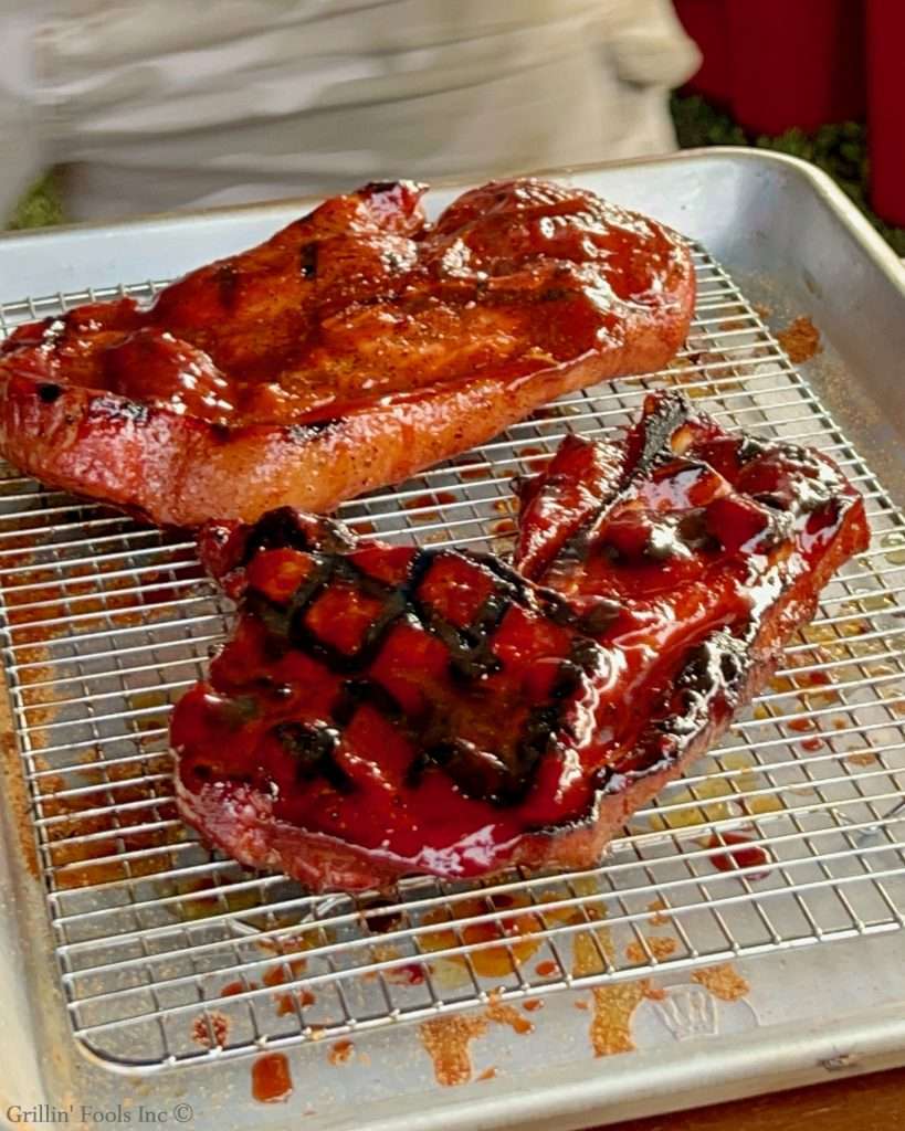 Glazed, competition pork steaks on a wire rack with a glossy red glaze and meat juices on a metal tray.