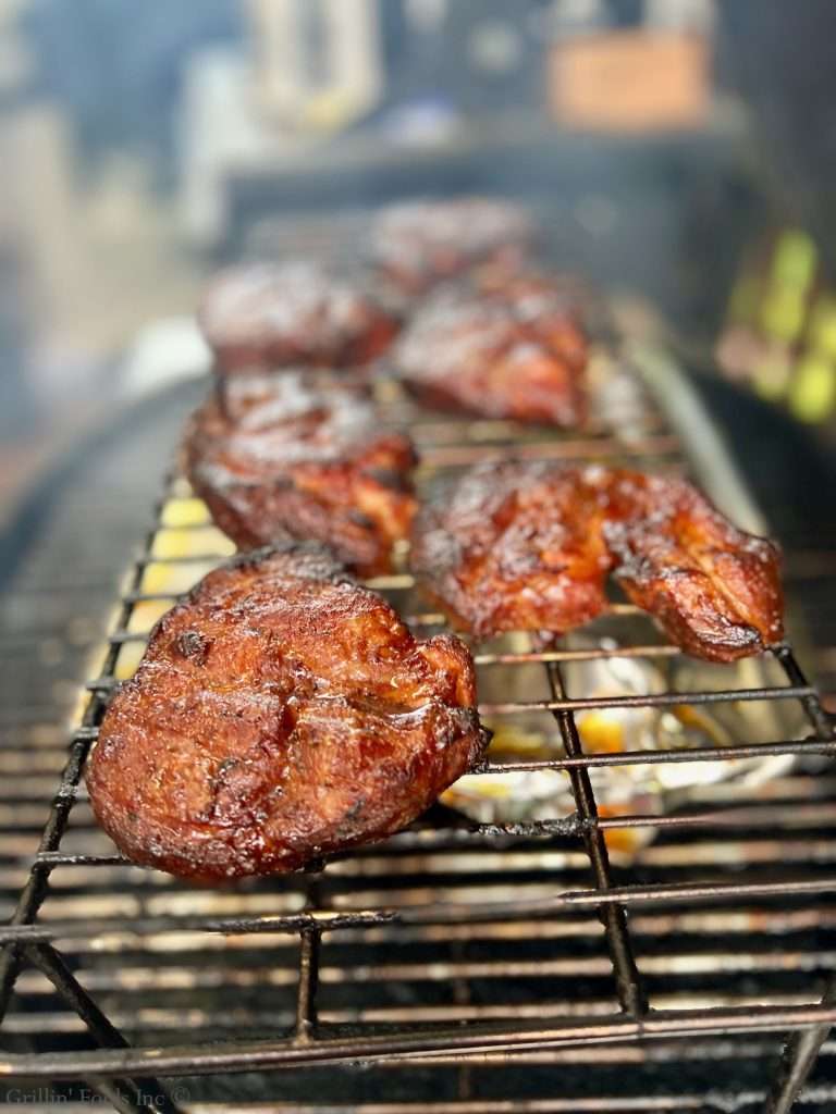 Smoked, half pork steaks on a grill rack, glazed and caramelized, cooking over hot coals in a smoky setting.
