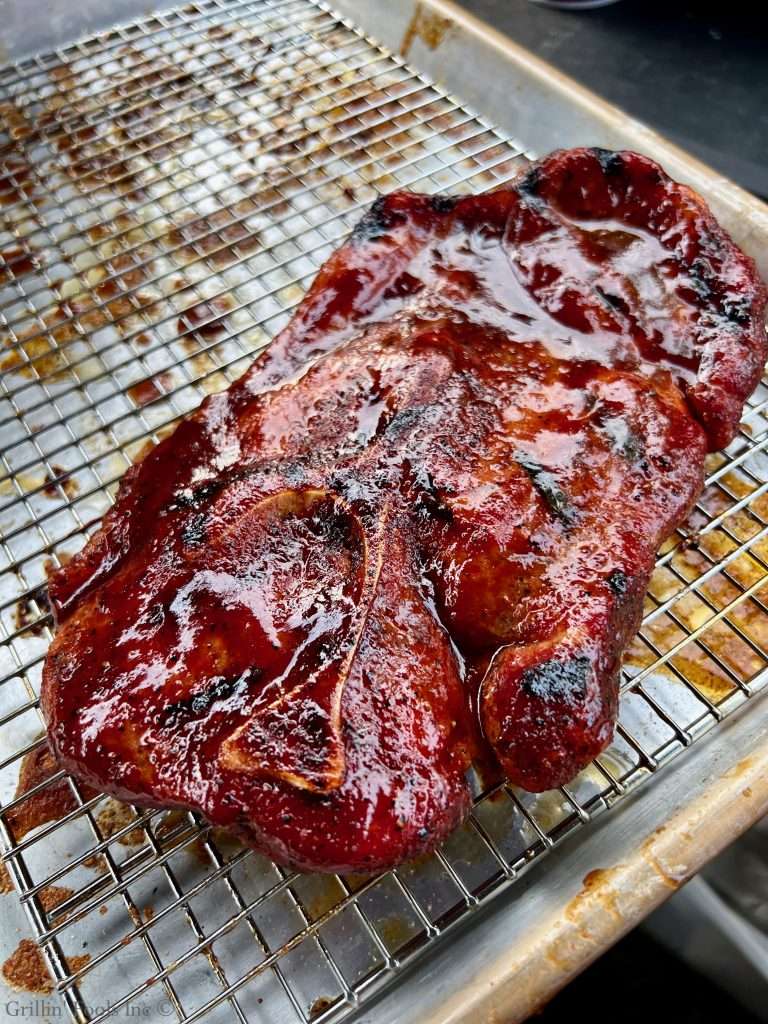 Barbecue pork steak on a wire rack with a glossy reddish glaze and charred edges, resting in a metal tray.