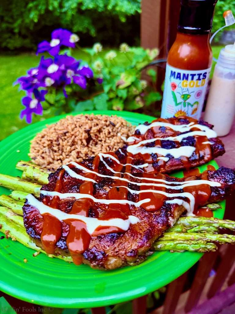 Plate of glazed barbecue pork steak with drizzle of white sauce, served with asparagus spears and brown rice outdoors near purple flowers and a hot sauce bottle.