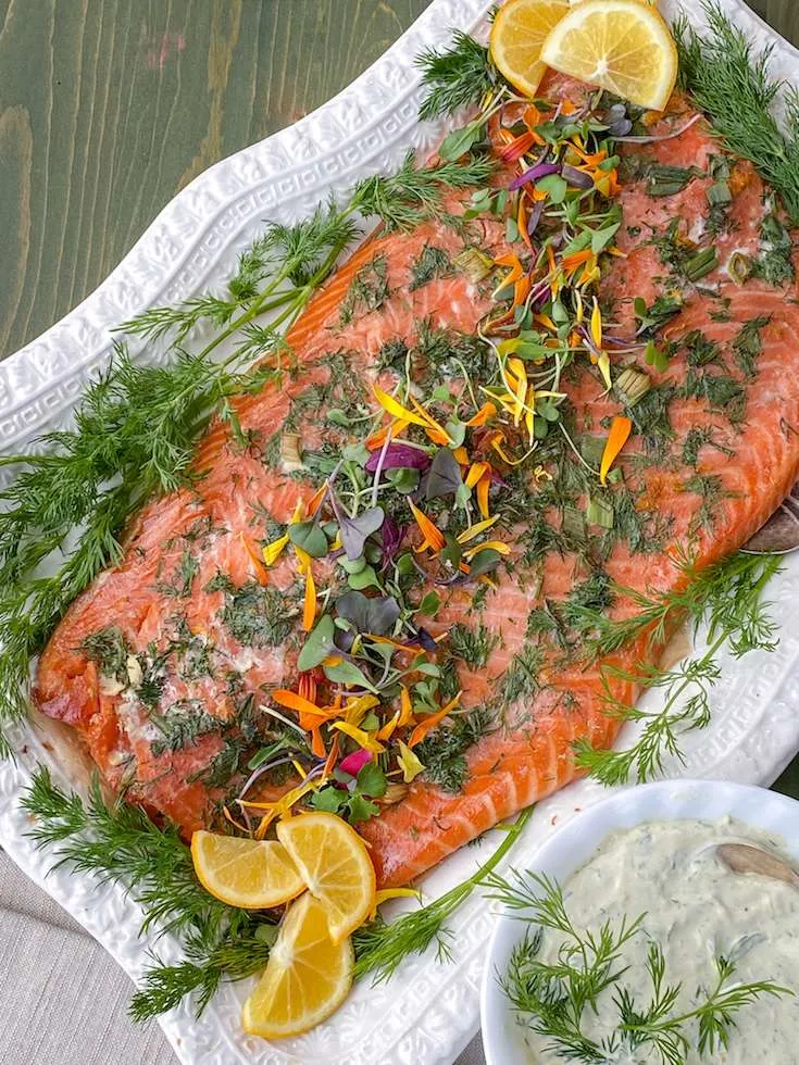 Herb-dressed salmon fillet on a platter with dill, edible flowers, lemon wedges, and a side herb sauce.