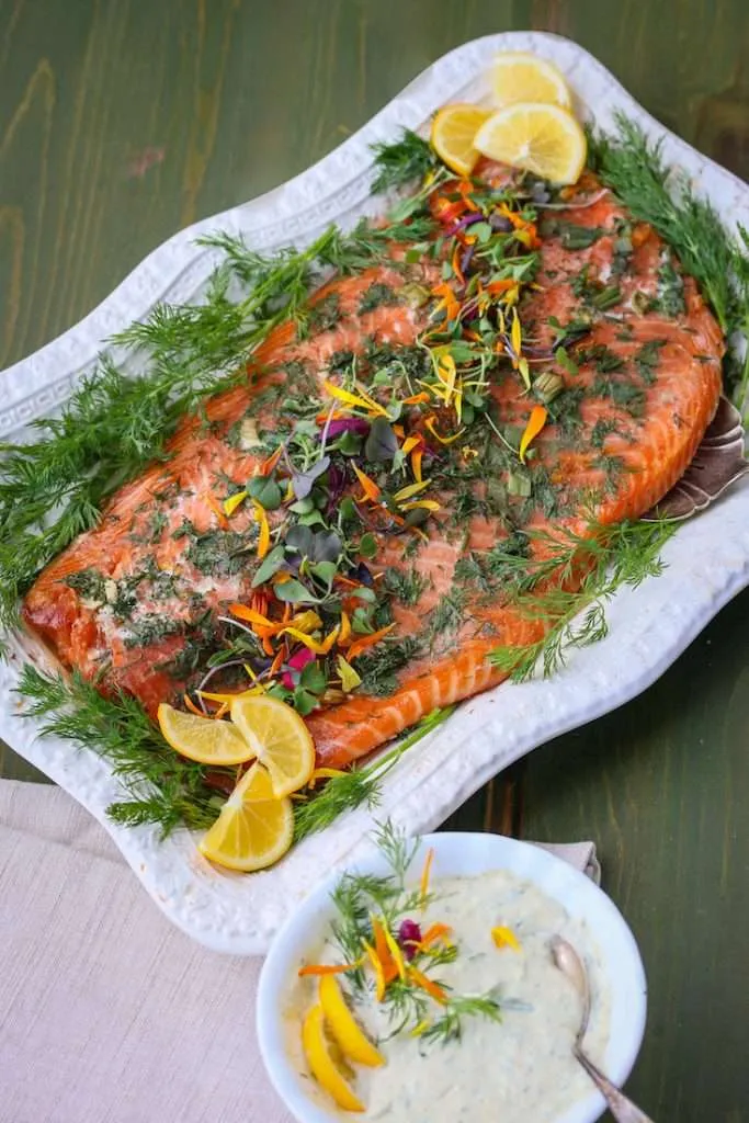Whole baked salmon on a white platter, garnished with dill, lemon wedges, and colorful edible flowers beside a creamy herb dip.