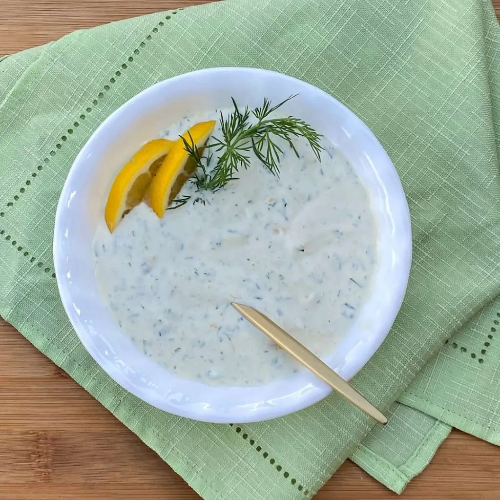 Creamy dill dip in a white bowl on sea foam green napkins with a couple slices of lemon, dill sprigs and a spoon