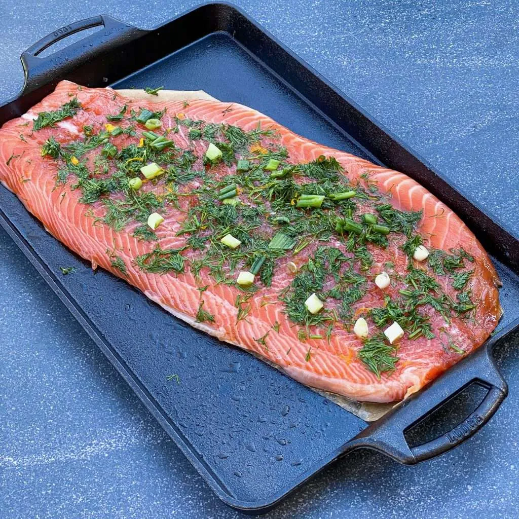 A side of brined salmon, topped with fresh dill and green onions, on a piece of salmon shaped parchment paper all in a cast iron, rimmed baking sheet.