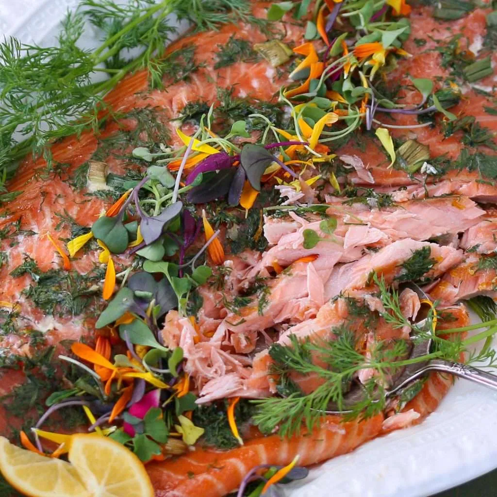 Salmon fillet topped with dill, herbs, and colorful edible flowers on a white plate with a lemon wedge nearby.