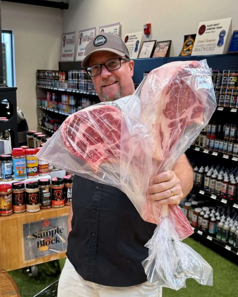 Smiling man in a shop holding 2 large raw steaks sealed in a clear plastic bag.