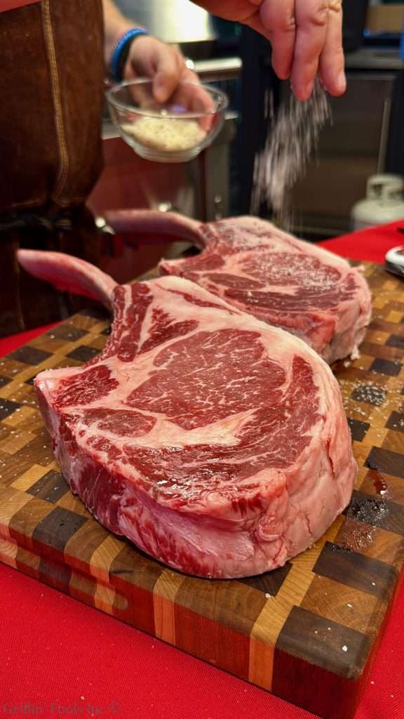 Two large raw ribeye steaks on a cutting board being seasoned with salt.