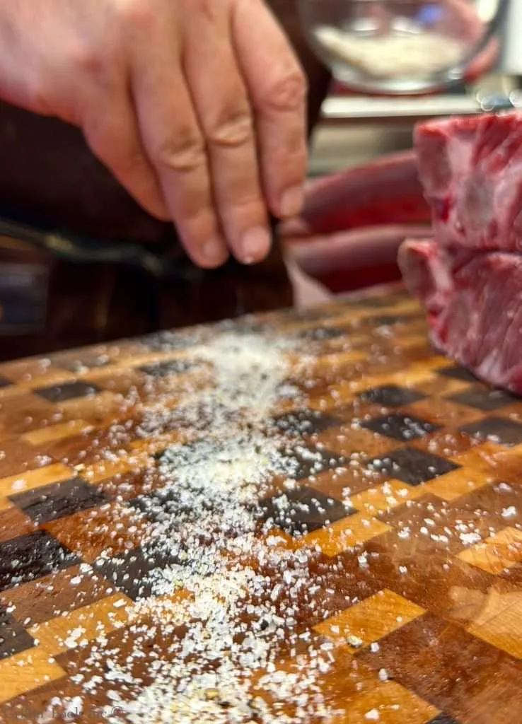 Close-up of a butcher's hand sprinkling coarse salt on a wooden cutting board with raw meat nearby.