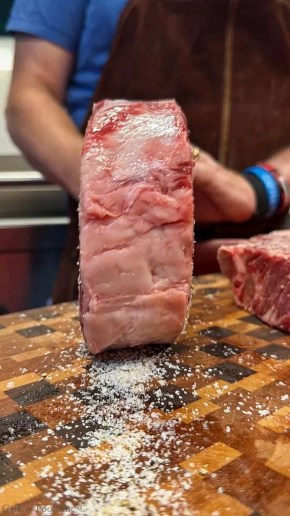 Thick raw beef steak on a wooden cutting board with coarse salt scattered around; person with blue shirt and brown apron in background.