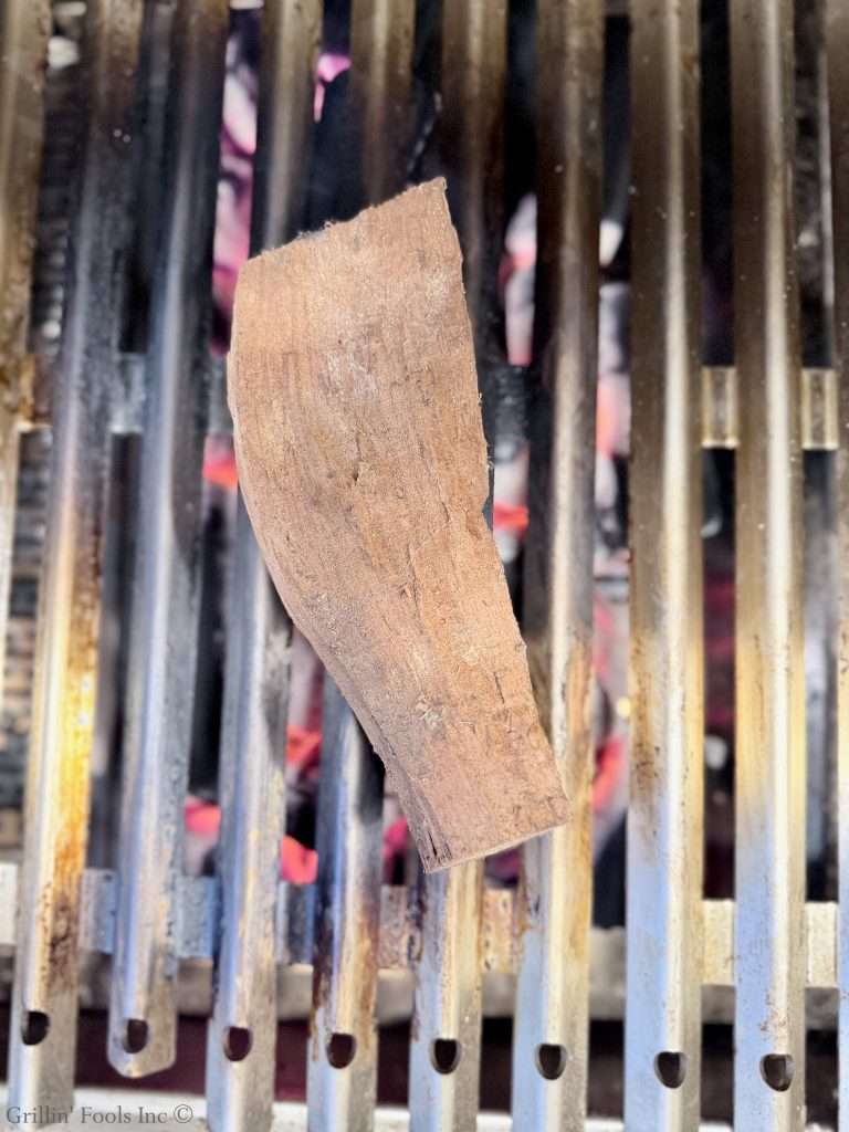 Close-up of a rough brown wooden plank resting on a a grill grate with lit charcoal glowing orange underneath