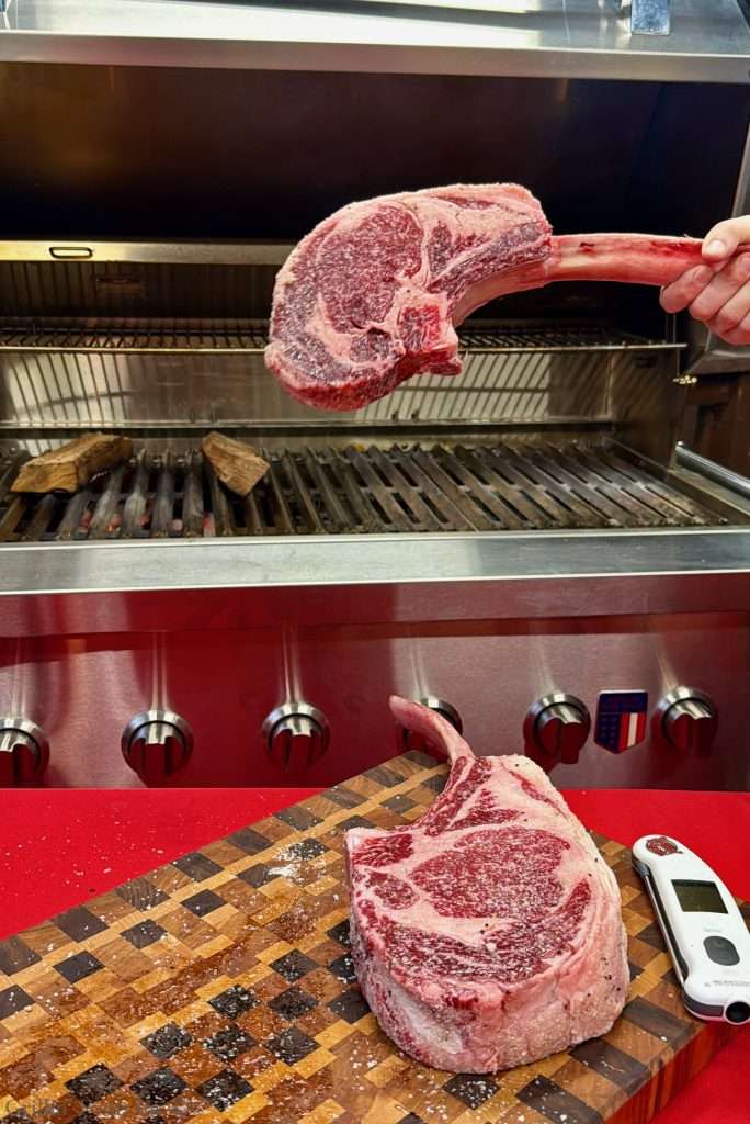 Raw bone-in ribeye steak with marbling on a wooden cutting board in a butcher shop, grill in the background.