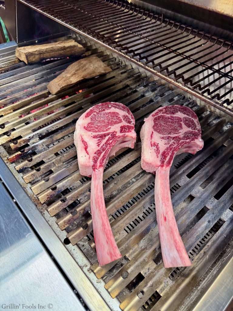 Two large bone-in ribeye steaks resting on a barbecue grill with wood chunks nearby (uncooked).