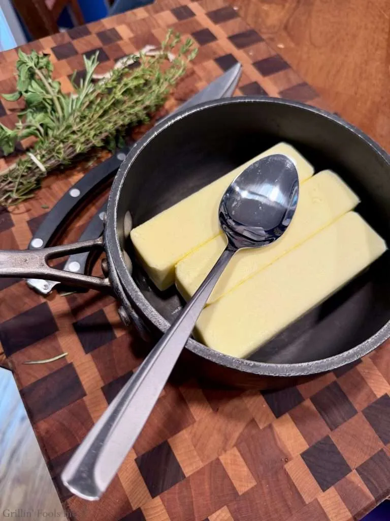 Three sticks of butter in a black saucepan with a spoon resting on top, on a wooden cutting board with fresh herbs nearby.