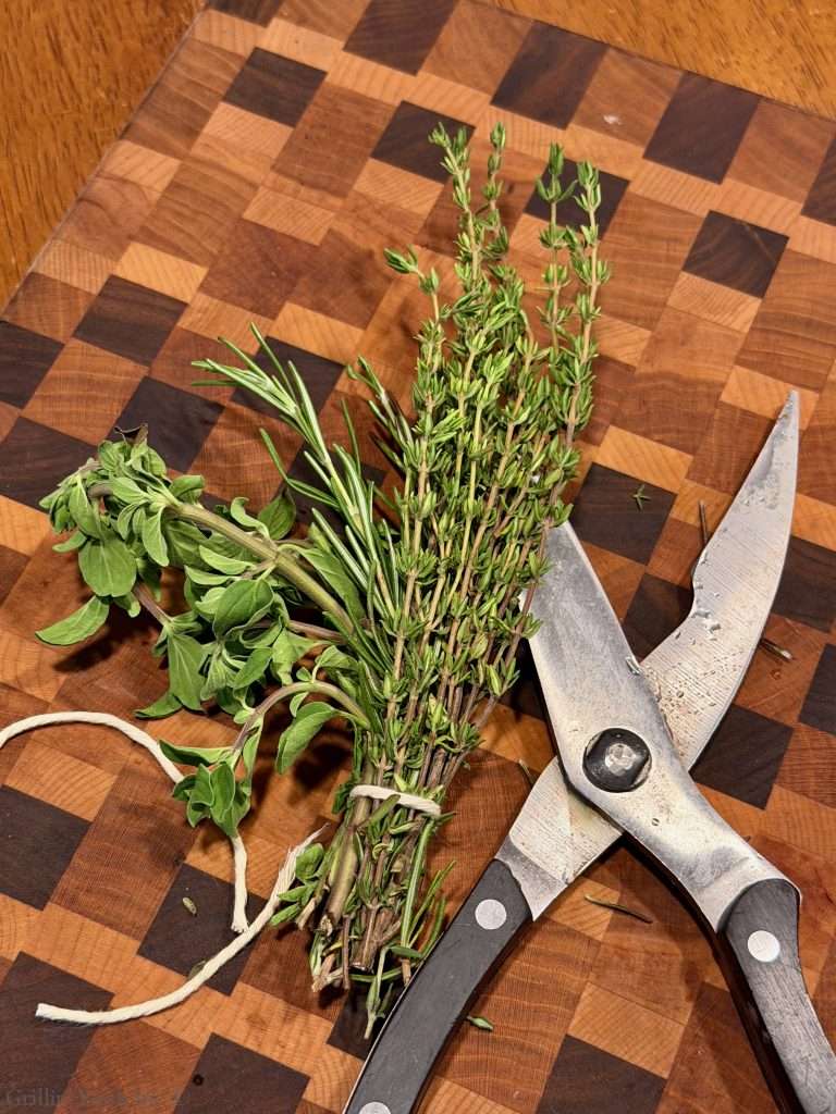 Bundle of fresh thyme and rosemary tied with string on a wooden cutting board, beside kitchen scissors.