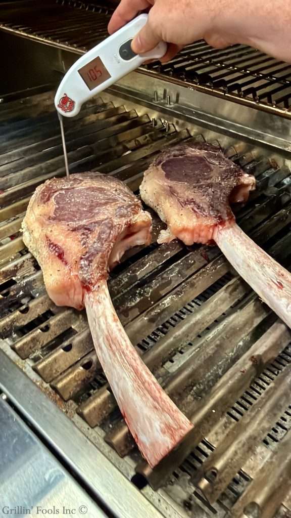 Hand holding a digital meat thermometer inserted into one of two tomahawk steaks on a hot grill, checking internal temperature (display shows 105°F).