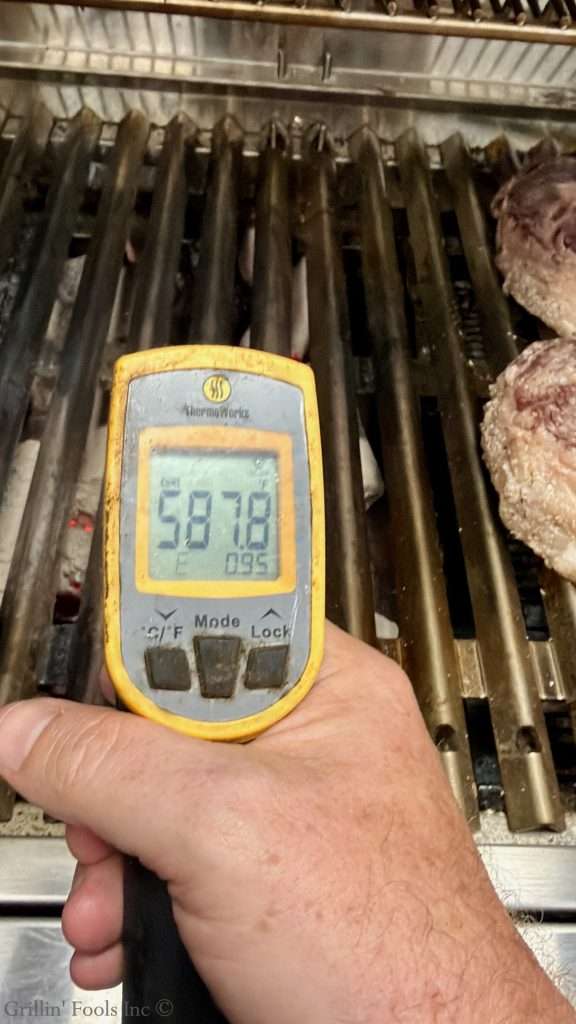 Handheld infrared thermometer over grill grates during cooking, with meat nearby. The display shows a temperature reading around 587°F.