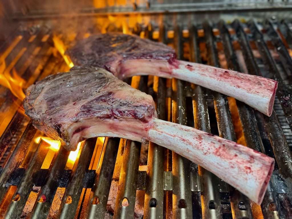 Bone-in ribeye steaks cooking on a grill over open flames, sizzling on the grate.