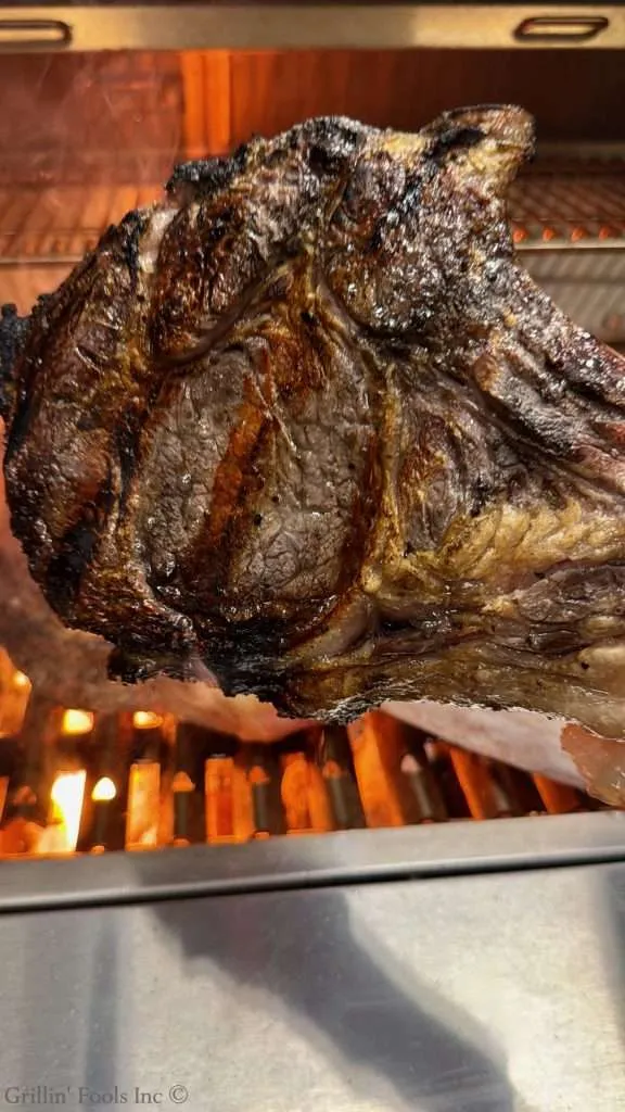 Large smoked and seared beef steak with a dark, charred crust being held by the bone over a hot grill