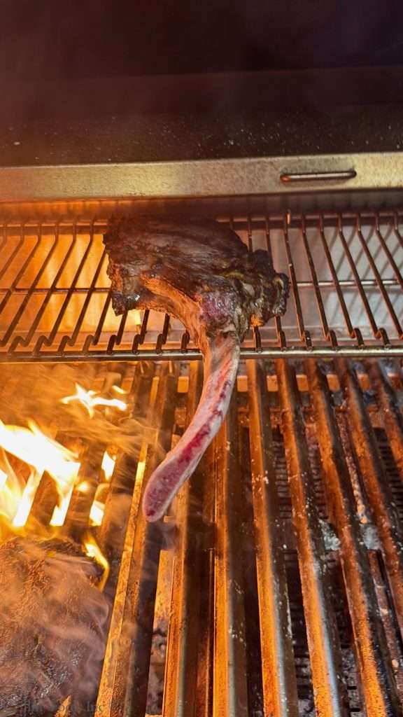 Tomahawk steak on a charcoal grill over open flames, meat browning on the grate.