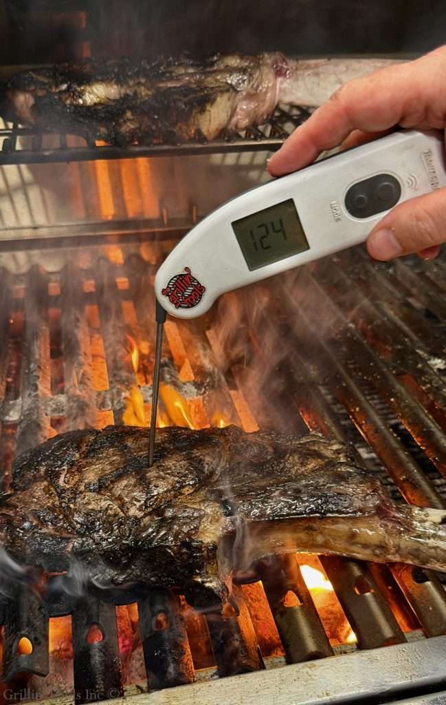 Person inserting a digital meat thermometer into a thick beef steak on a hot grill, display reading 124°F (literally thermometer showing).