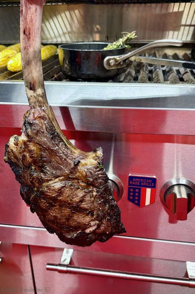 A large reverse sear steak held by the bone, with a black cooking pot on the grate and corn on the cob visible in the background.