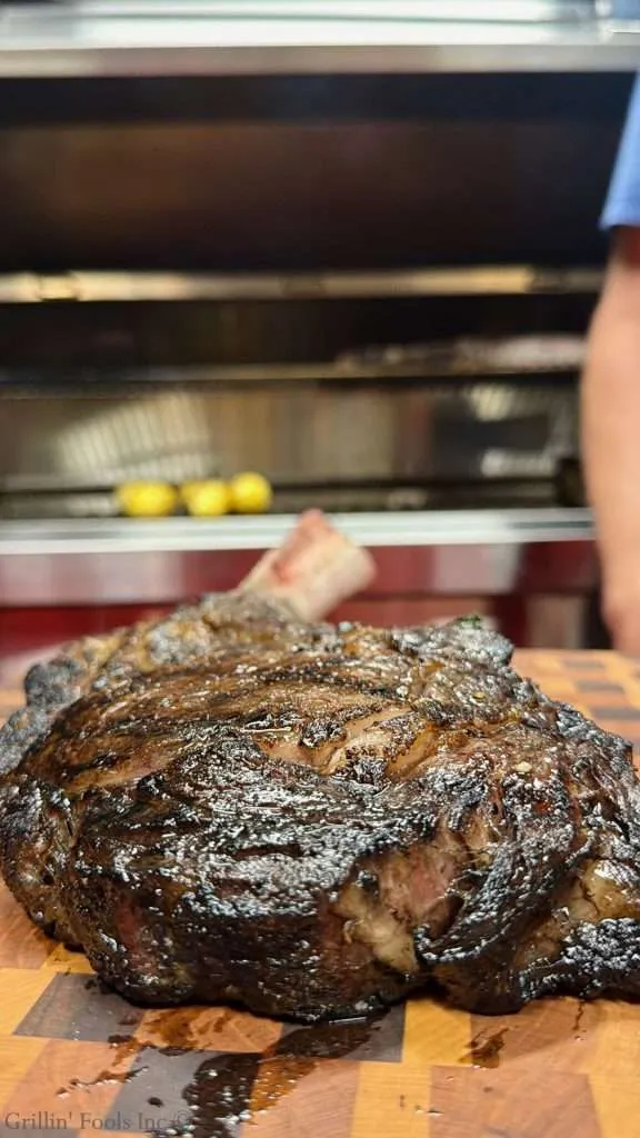 Large reverse sear tomahawk steak with bone on a checkered cutting board, glossy juices, in a kitchen with a grill in the background.