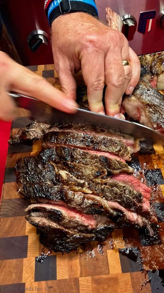 Hands slicing a large ribeye steak on a wooden cutting board with a sharp knife.
