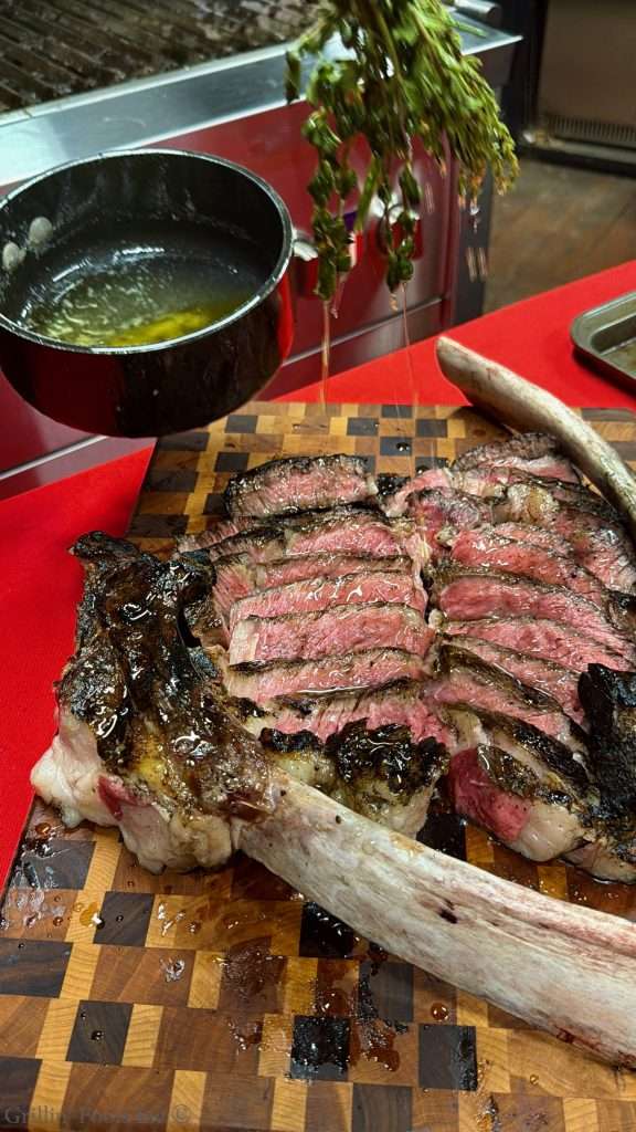 Sliced medium-rare ribeye steak arranged on a wooden cutting board with a bone nearby and herbs hanging above, while clarified butter is drizzled over the top with a fresh herb mop