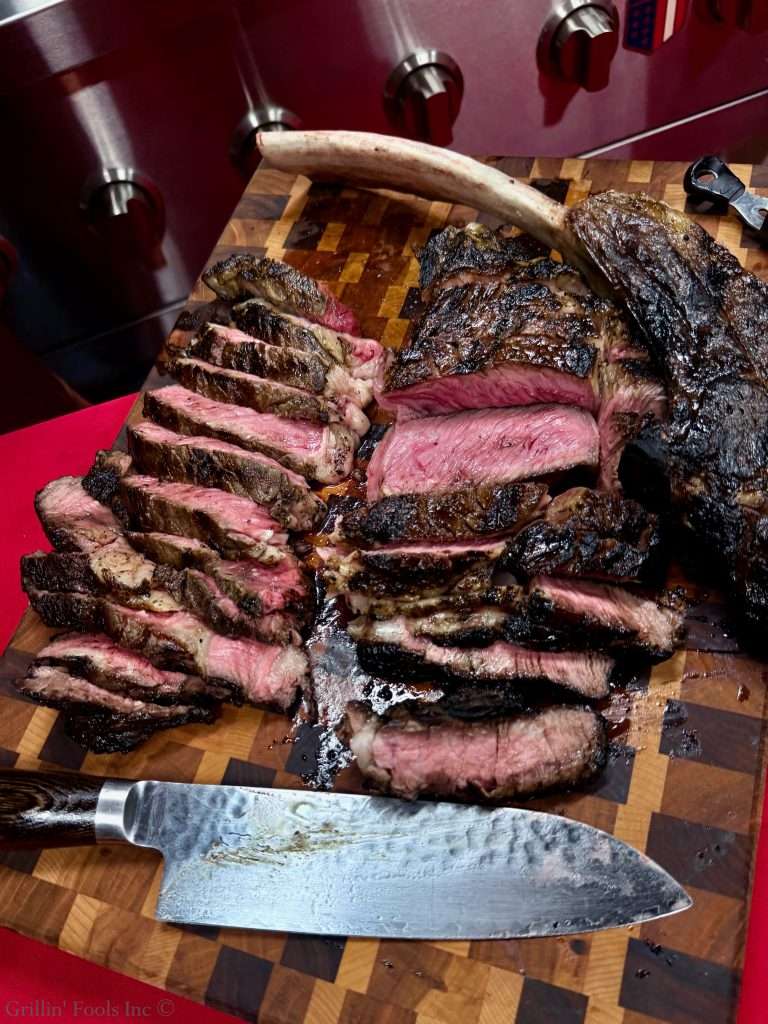 Sliced ribeye on a wooden cutting board, with a large bone bone-in piece resting on top and juices around it from the tomahawk steak recipe