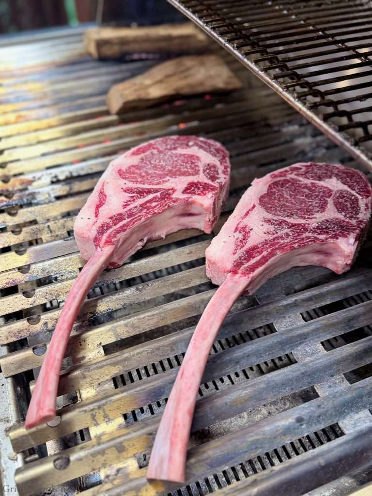 Two raw bone-in ribeye steaks with prominent marbling on a metal grill, ready to cook outdoors for a tomahawk steak recipe