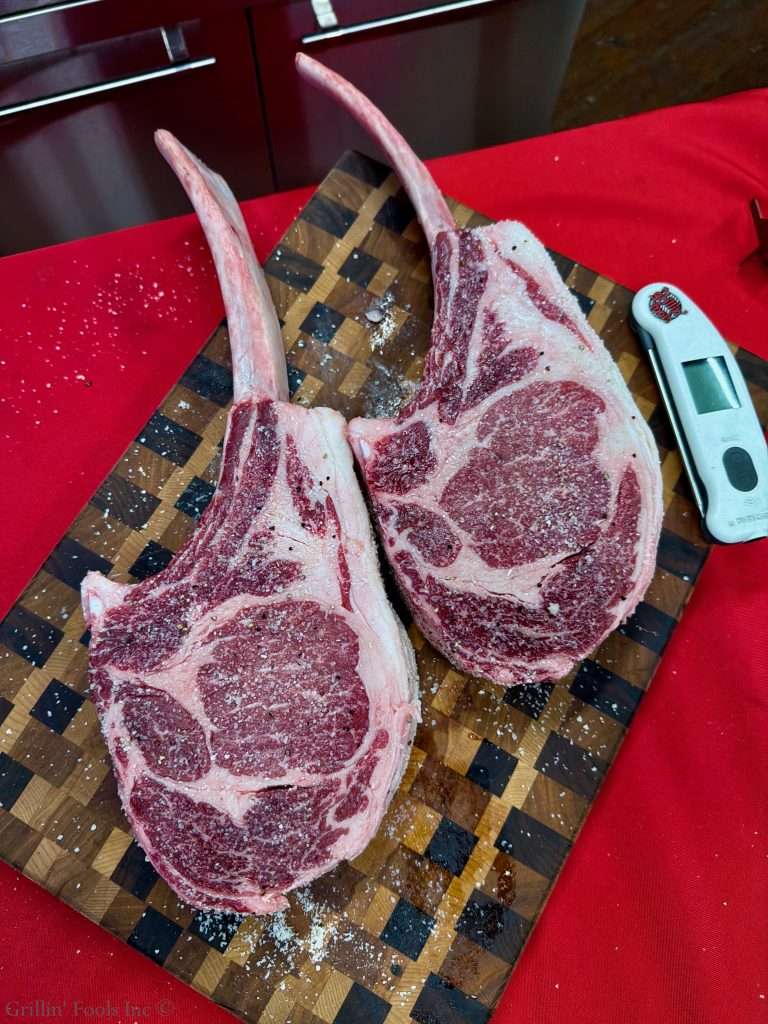 Two large raw tomahawk ribeye steaks with long bones on a wooden cutting board, sprinkled with salt, on a red surface, with a digital meat thermometer nearby ready for a photoshoot for this tomahawk steak recipe