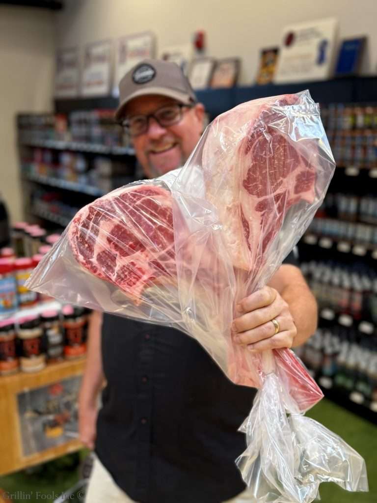 Shop attendant proudly holds a large bone-in ribeye wrapped in plastic in a grocery store.