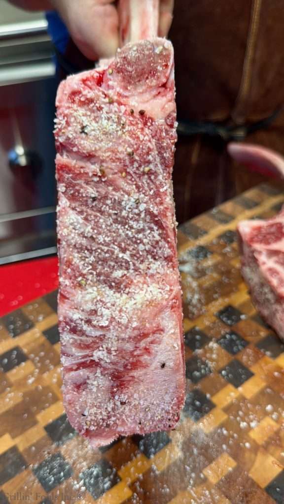 Hand holding a salt-crusted raw beef rib steak, with coarse salt and pepper visible on the surface.