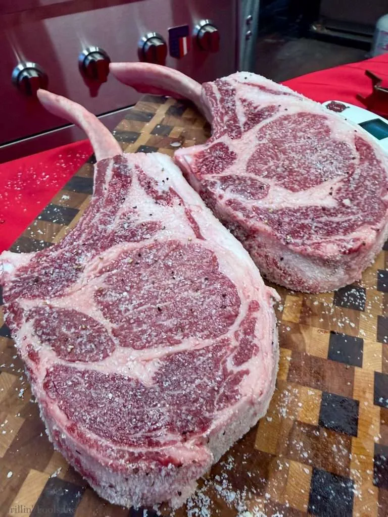 Two large raw tomahawk steaks on a salt-dusted cutting board, bone handles visible, with a stainless grill in the background.