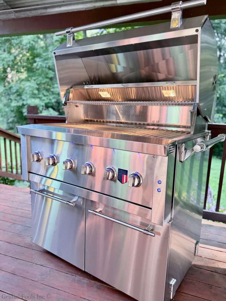 Brand new American Made Grills, Muscle Grill on a deck, with the lid open and the interior lights on inside the grill, with lots of green foliage in the background