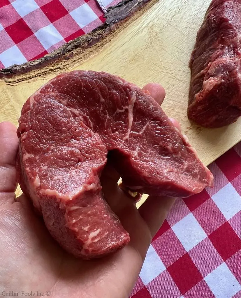 Raw coulotte steak bent over like picanha, held in a hand over a wooden cutting board with a red-and-white checkered tablecloth in the background.