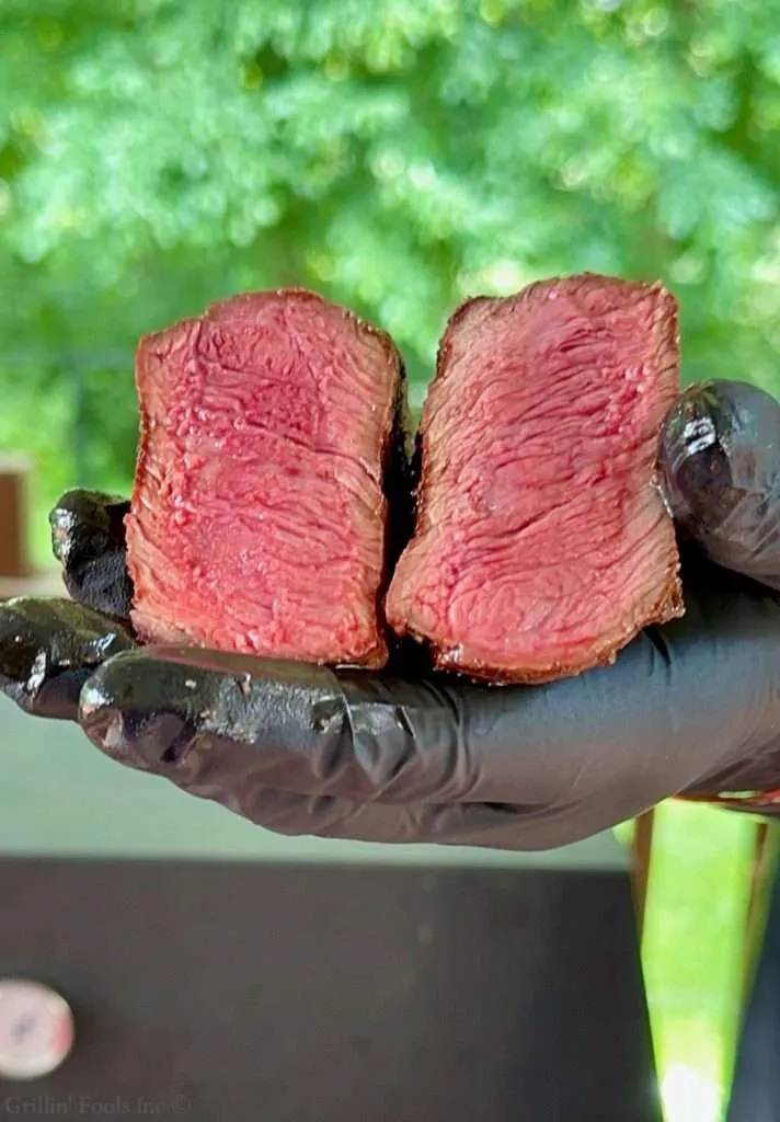 Close-up of two slices of medium-rare rump steak held by gloved hands with a green garden background