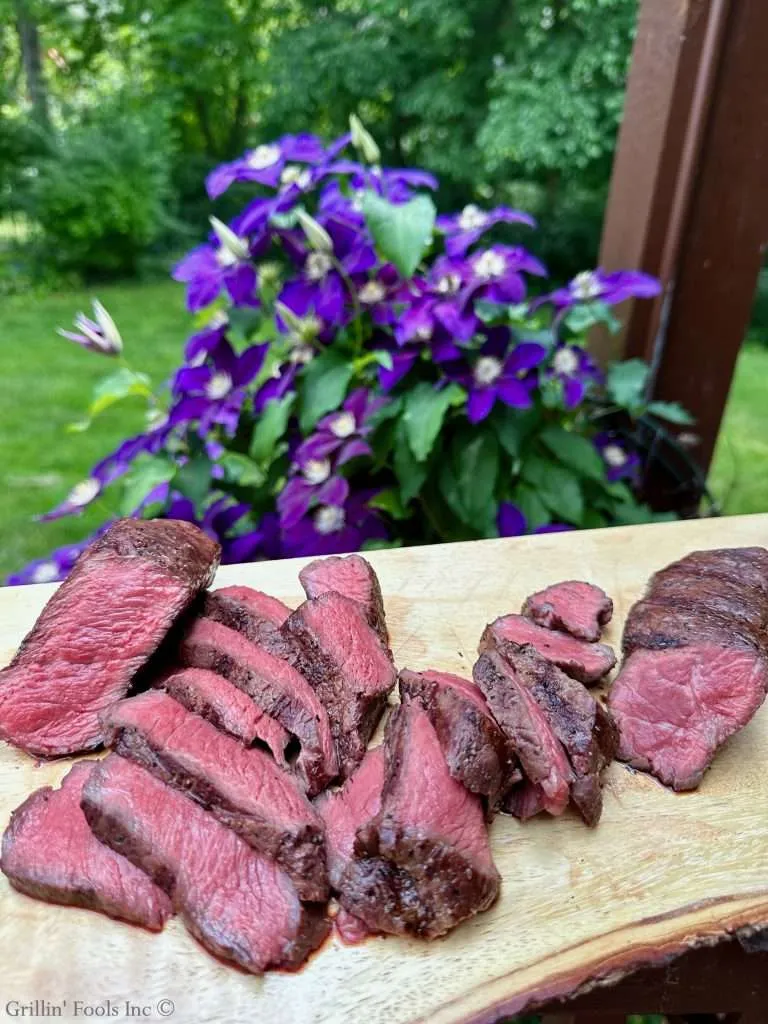 Slices of medium-rare grilled coulotte sirloin steak arranged on a wooden cutting board, outdoors with purple flowers in the background.