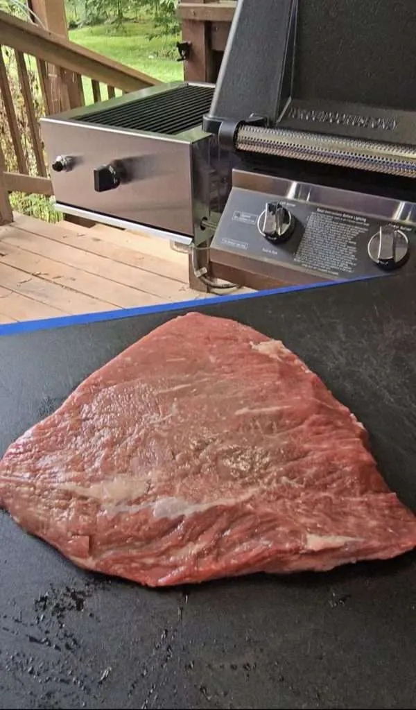 Raw rump cap on a dark cutting board with a stainless steel grill in the background on a wooden deck.