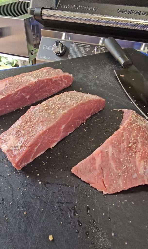 Raw sirloin coulotte steaks seasoned with salt, pepper and garlic on a black cutting board, with a knife visible in the background.