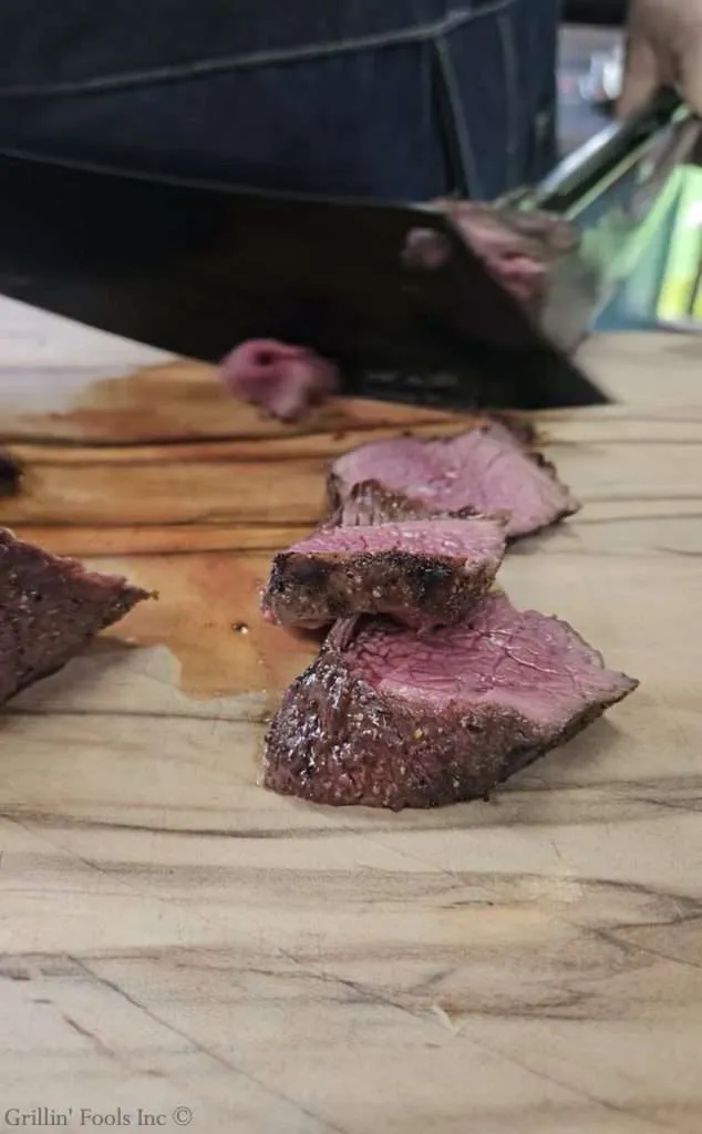 Sliced grilled coulotte steak on a wooden cutting board with a chef in the background.